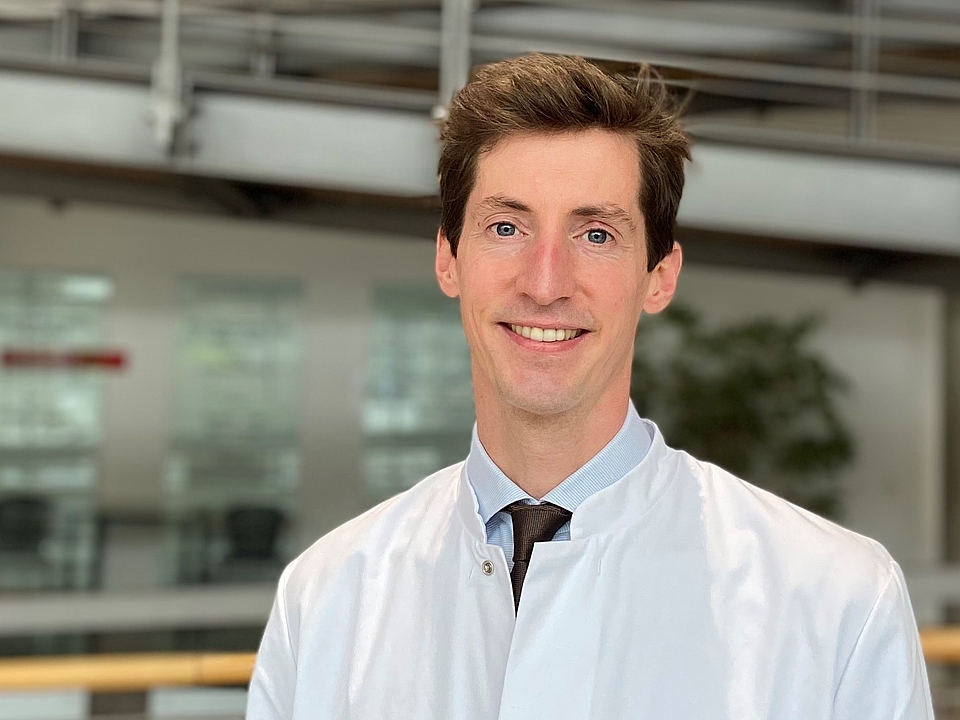  Ein junger Mann mit kurzen, dunklen Haaren und einem weißen Arztkittel lächelt in die Kamera. Er steht in einem hellen, modernen Gebäude mit einer Rampe und Geländern im Hintergrund.