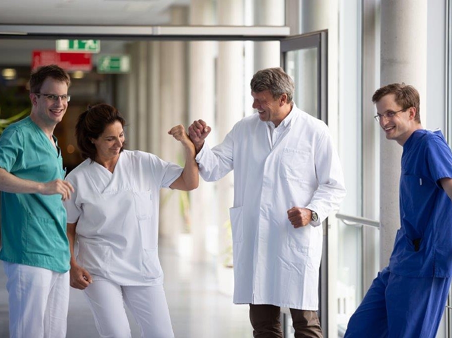 Vier lächelnde Ärzte des Darmkrebszentrums des Krankenhauses Agatharied stehen in einem hellen Flur. Eine Ärztin und ein Arzt machen einen freundlichen Faust-Check. Ein weiterer junger Arzt lehnt an einem Geländer.