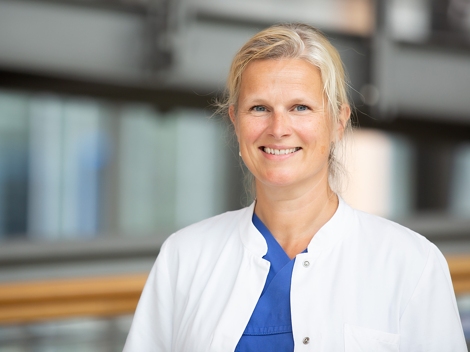 Eine lächelnde Frau mittleren Alters mit blondem Haar, die einen weißen Arztkittel über blauer medizinischer Kleidung trägt und in die Kamera blickt.