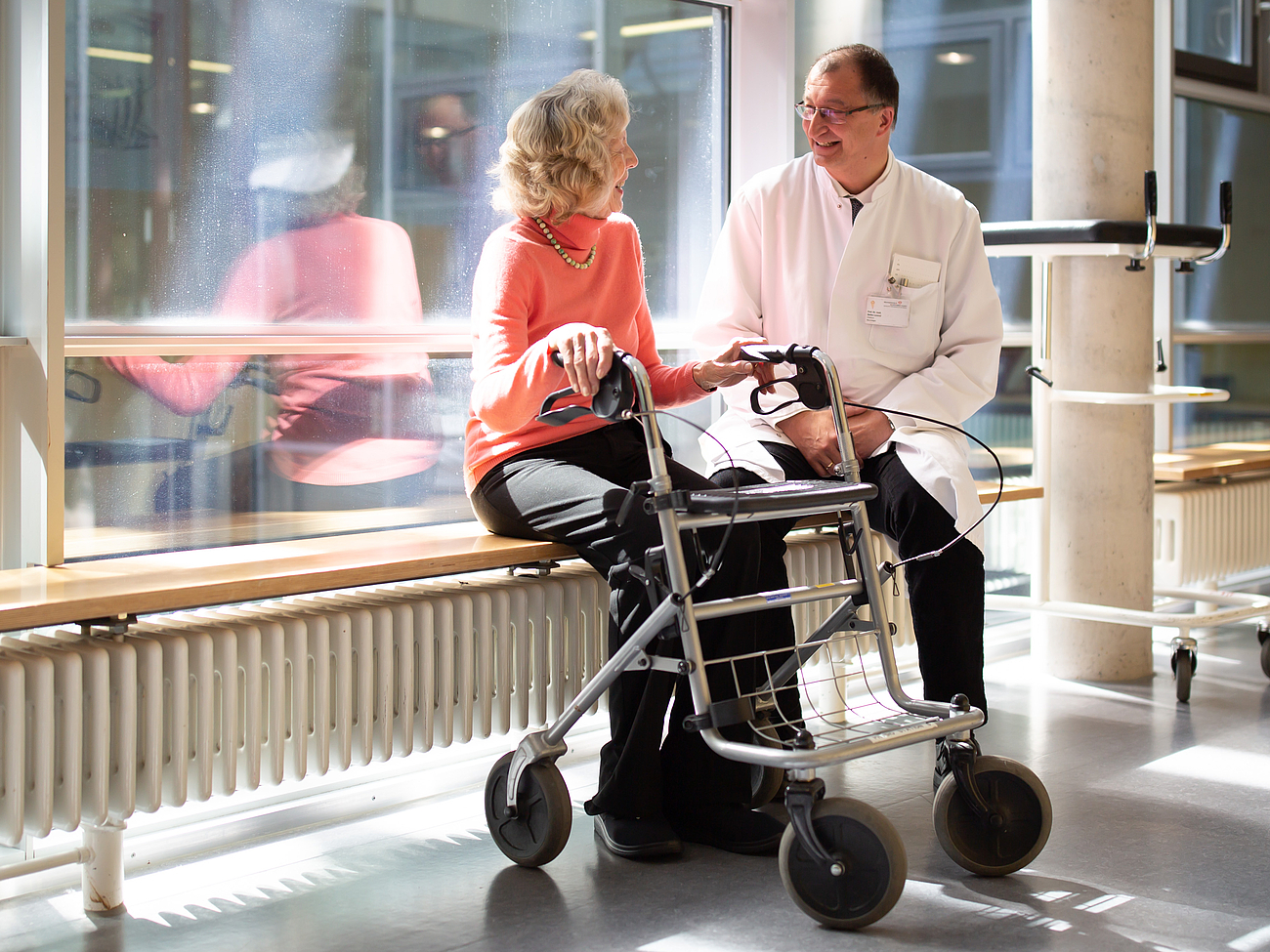  Ein Arzt in weißem Kittel sitzt auf einer Bank neben einer älteren Patientin, die ebenfalls auf der Bank sitzt und einen Rollator vor sich hat. Sie unterhalten sich in einem hellen Flur mit großen Fenstern.