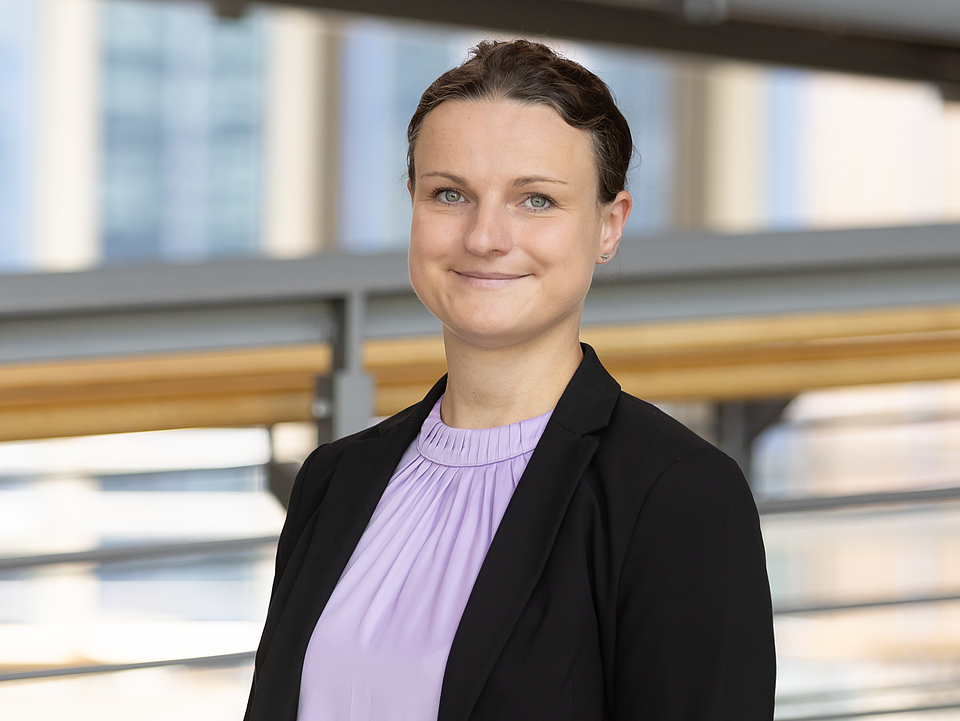 Porträt Lehmann Eine junge Frau mit zusammengebundenen, dunklen Haaren und einem leichten Lächeln trägt eine lilafarbene Bluse und einen schwarzen Blazer. Sie steht im Foyer des Krankenhauses mit hellen Geländern und Fenstern im Hintergrund.
