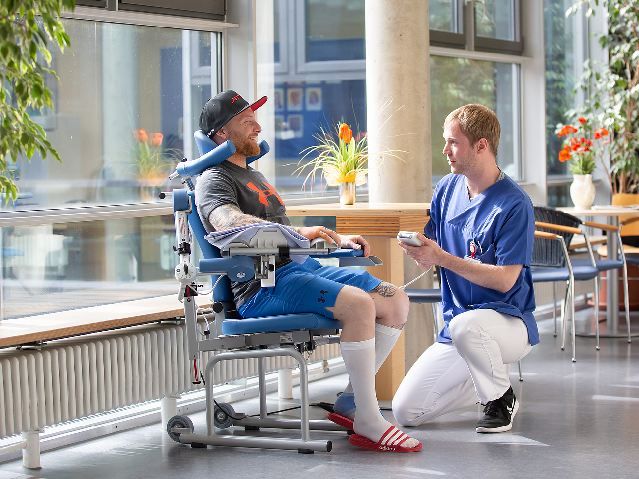 Patient und Pfleger im Flur Ein Patient sitz auf einem Stuhl im Flur und vor ihm kniet ein Pfleger und hilft ihm.